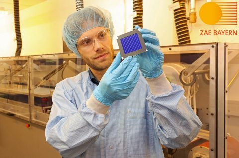 Ein Mann in Laborkittel, -handschuhen und -haube hät ein großes Solarmodul hoch