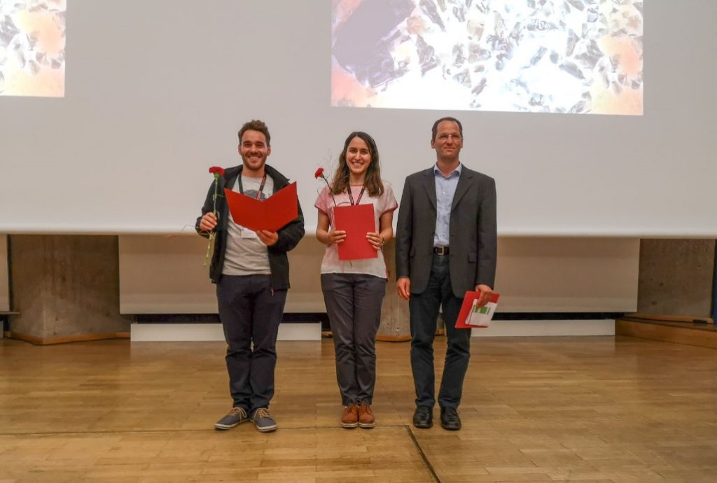 Drei Personen stehen nebeneinander, rechts und links je ein Mann, in der Mitte eine Frau. Die beiden links halten eine rote Mappe sowie je eine rote Rose in den Händen.